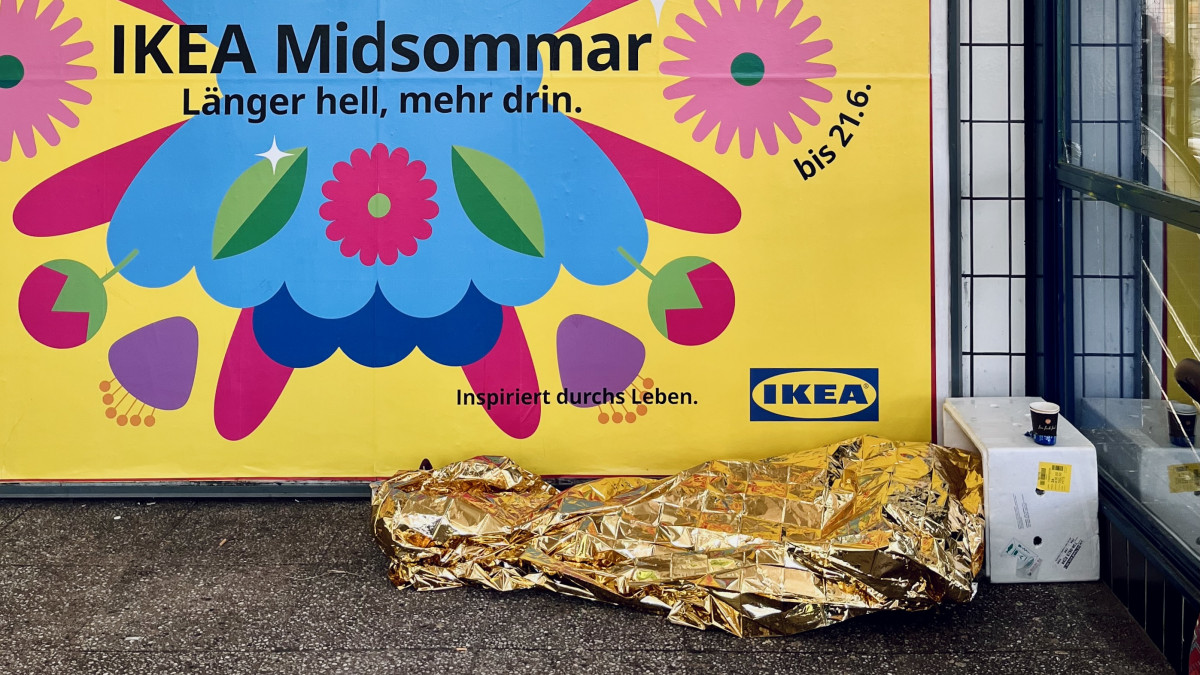 Person mit goldener Rettungsdecke vor einem IKEA-Werbeplakat im öffentlichen Raum