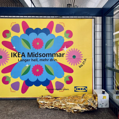 Person mit goldener Rettungsdecke vor einem IKEA-Werbeplakat im &ouml;ffentlichen Raum